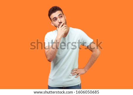Need to think. Portrait of thoughtful brunette man with beard in white t-shirt holding his chin and pondering idea, confused not sure about solution. indoor studio shot isolated on orange background Royalty-Free Stock Photo #1606050685