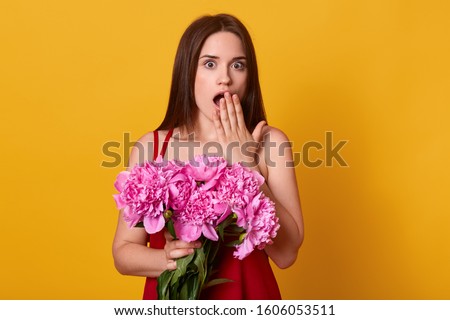 Studio shot of surprised young woman covering her opened mouth with hber palm, looks in amazement, holds bunch of pink peonies, posing isolated over yellow studio wall. St. Valentines Day concept.