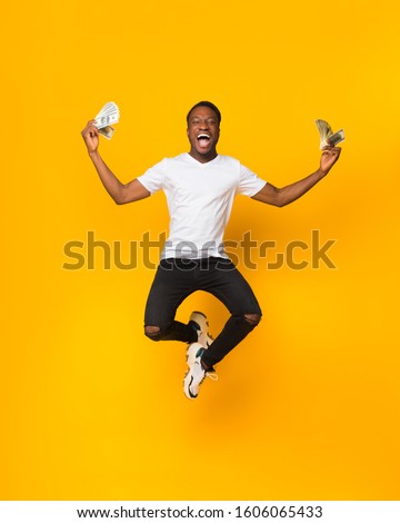 Excited African American Guy Jumping Holding Bunch Of Money Celebrating Profit And Success Over Yellow Studio Background. Big Luck Concept