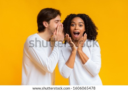 Young man sharing secret with his shocked black girlfriend, gossiping together over yellow background with free space