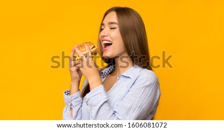 Cheat meal day. Hungry young woman eating burger standing in studio on yellow background. Panorama