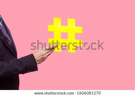 Closeup of male hand in stylish black tuxedo holding large yellow hashtag symbol, popularity and promotion in social media, viral content, trending. indoor studio shot isolated on pink background