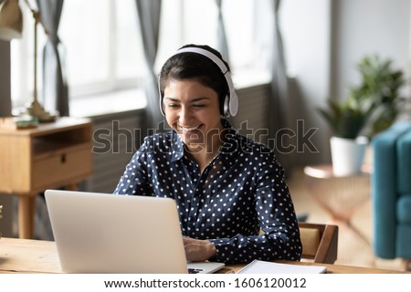 Happy indian young girl student wear headphone watch webinar listen online course communicate by conference video call e learn language in app laugh study with teacher lesson look at laptop at home Royalty-Free Stock Photo #1606120012