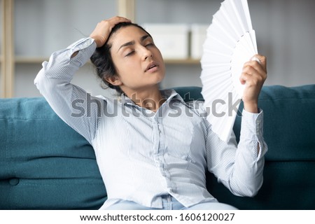 Tired sweaty young indian woman holding using hand fan sit on couch in uncomfortable hot summer weather suffer without air conditioner cooling feel hot at home apartment, overheating concept Royalty-Free Stock Photo #1606120027