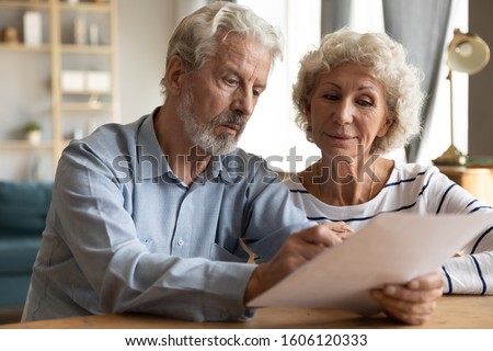 Focused elderly 70s married couple sit at table at home, husband holding papers documents spouses manage financial statement analysing medical insurance terms, bank account balance of family concept Royalty-Free Stock Photo #1606120333