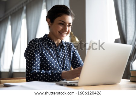 Smiling indian girl student professional employee typing on laptop sit at home office table, happy hindu woman studying e learning online software using technology app for work education concept Royalty-Free Stock Photo #1606120426