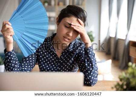 Tired overheated young indian woman hold wave fan suffer from heat sweating indoor work on laptop at home office, annoyed girl feel uncomfortable hot summer weather problem no air conditioner concept Royalty-Free Stock Photo #1606120447