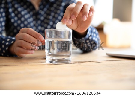 Young sick ill woman hold dissolving effervescent aspirin pill drop tablet into glass of sparkling water take painkiller medicine to relieve headache pain, healthcare medicament concept close up view