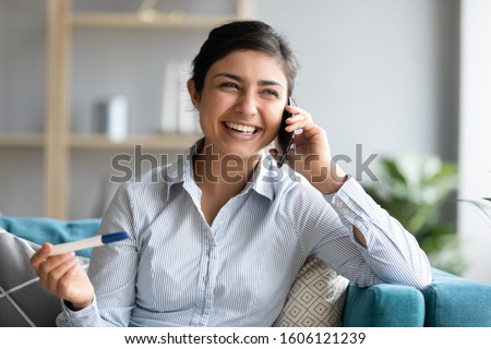 Excited pregnant young indian woman wife holding positive pregnancy test result calling husband talking on phone, overjoyed expectant mother make call share great news expect baby sit on sofa at home