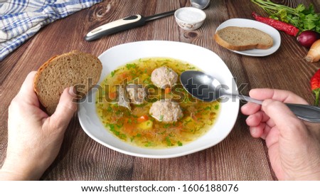 Soup with vegetables and meatballs in a white plate on a wooden table. Female hands holding a spoon and a slice of dark bread