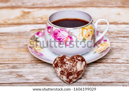 A white porcelain Cup on a saucer with a delicate pattern of pink and yellow rose buds with instant coffee on a wooden background with a heart-shaped chocolate gingerbread.