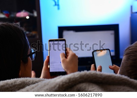 friendship, people, technology and entertainment concept - A mother and son holds a smartphones in theirs hands using a DLNA in the living room in front of the TV, enjoying numerous content over. Royalty-Free Stock Photo #1606217890