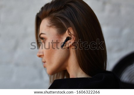 Young brown hair street fashion woman in black hoodie with opened neck turned back and uses to talk or listen music wireless earphones in ear in loft interior with white brick wall Royalty-Free Stock Photo #1606322266