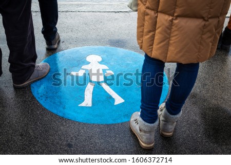 People waiting at pedestrian crosswalk in city street. Road sign on asphalt road. Human legs crossing street