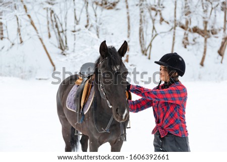 A beautiful brunette girl in a plaid black-and-white shirt walks with a big black horse in a snowy park. A woman in a helmet for riding walks in a snowy forest with a thoroughbred horse.