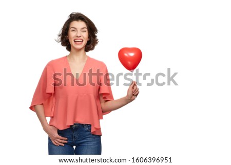 A beautiful brunette Girl with a wide smile in jeans and a pink jacket on a white background holding red balloons in the form of a heart