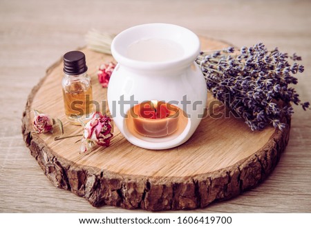 Vintage style picture of white ceramic candle aroma oil lamp with essential oil bottle and dry flower petals on natural pine wood disc, dry background with copy space.