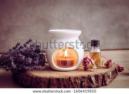 Vintage style picture of white ceramic candle aroma oil lamp with essential oil bottle and dry flower petals on natural pine wood disc, dry background with copy space. Royalty-Free Stock Photo #1606419850
