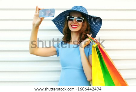 Portrait happy smiling woman taking selfie picture by smartphone with shopping bags wearing blue dress, summer straw round hat and sunglasses over white wall background