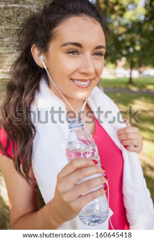 Happy young sporty woman leaning against tree holding a bottle of water