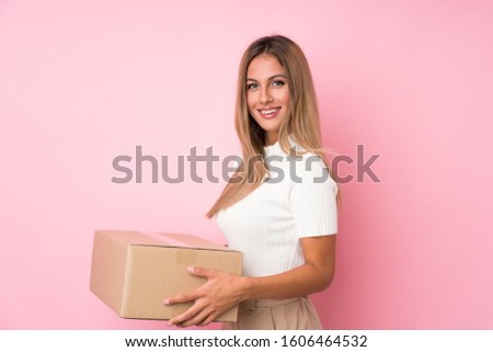 Young blonde woman over isolated pink background holding a box to move it to another site