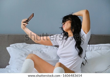 Beautiful brunette woman sitting in white bed, relaxing at home, taking selfie, chatting on social networks, female beauty blogger taking photos and videos for blog