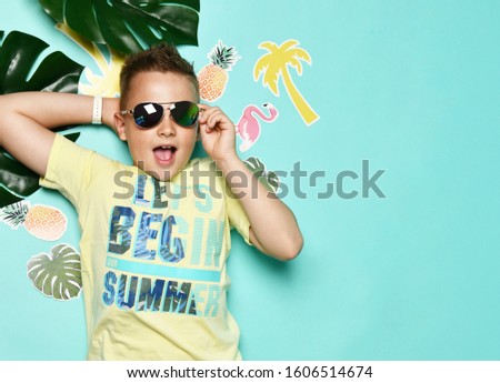 boy is  lying on the floor in a yellow T-shirt and sunglasses a happy smile with paper palms and flamingos and palm trees on a blue background. The concept of summer holidays, vacation at the resort