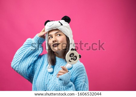 Funny young girl standing with yummy panda-lollipop in her hand and a hat on his head on a pink background.