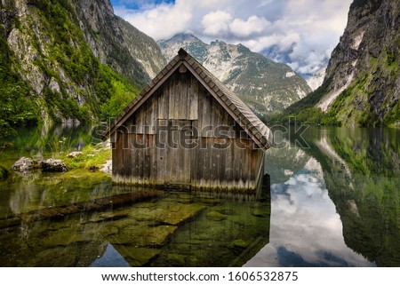 A wooden cabin, used as a boathouse, in the clear water of Obersee Lake surrounded by high Alps mountains in Schoenau am Koenigssee in Bavaria, Germany at the border with Austria.   Royalty-Free Stock Photo #1606532875
