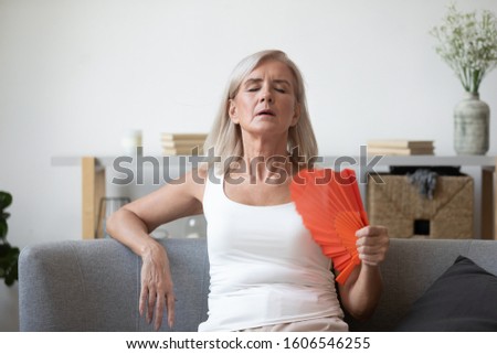 Unwell elderly female retiree sit on couch feel dehydrated tired of heat use hand waver to cool down, overheated stressed elderly woman suffer from heatstroke in living room with no air conditioner Royalty-Free Stock Photo #1606546255