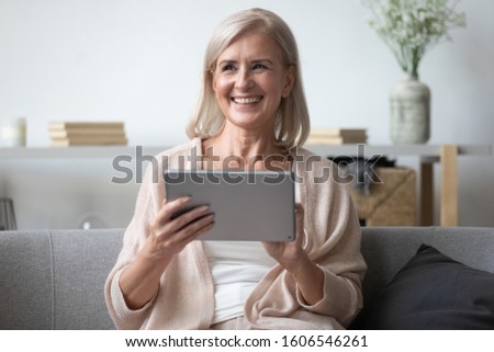 Happy senior woman sit on cozy couch relax in living room at home look in distance thinking using tablet watching movie, smiling mature female dreaming rest on sofa with electronic device in hands