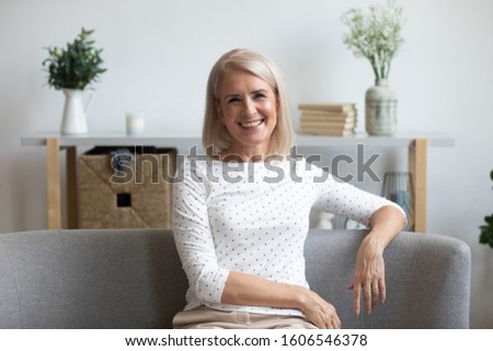 Close up headshot portrait of smiling positive elderly woman sit on couch in living room look at camera, happy middle-aged female pensioner rest relax on cozy sofa at home posing for picture indoors