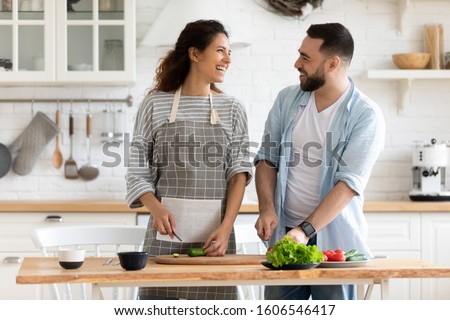 30s spouses looking at each other preparing healthy salad standing on cozy kitchen, holding knifes cutting fresh vegetables on wooden board enjoy time together and cooking process on weekend at home Royalty-Free Stock Photo #1606546417