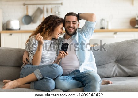 Cheerful married couple resting on couch enjoy lazy weekend together at home having fun using online amusing apps, wife holding smart phone spouses laughing on funny video, website with pranks concept Royalty-Free Stock Photo #1606546429