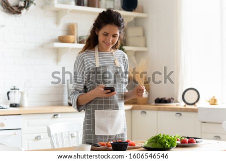 Woman wearing apron standing on domestic kitchen table full of fresh vegetables, housewife holds phone using cooking apps websites search recipes, chatting with friend distracted from food preparation