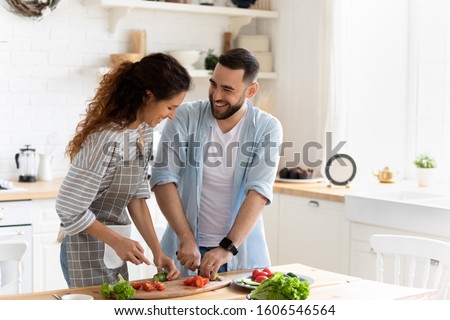 Happy couple standing in kitchen at home preparing together yummy dinner on first dating, spouses chatting enjoy warm conversation and cooking process, caring for health, eating fresh vegetable salad Royalty-Free Stock Photo #1606546564