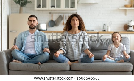 Horizontal banner serene couple and little daughter sitting on sofa in lotus position closed eyes do meditation breathing technique in living room, keep calm, healthy life habits and lifestyle concept Royalty-Free Stock Photo #1606546567