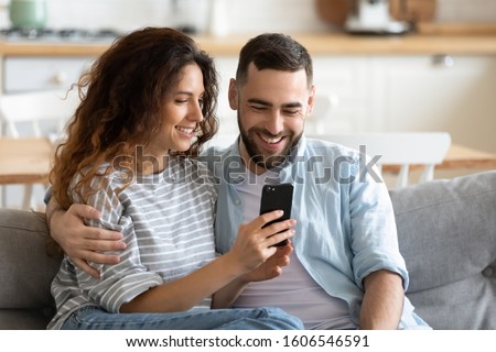 Couple in love resting on comfy couch having fun using smart phone apps, enjoy distant video call, wife showing interesting website to husband, choosing services goods on-line e-commerce usage concept Royalty-Free Stock Photo #1606546591