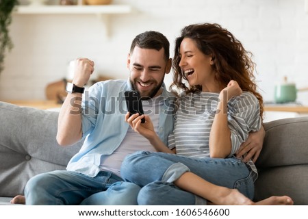 Excited overjoyed couple resting on couch holding smart phone celebrating on-line lottery win, bid betting victory moment, unbelievable opportunity or invitation, internet sale, getting prize concept Royalty-Free Stock Photo #1606546600