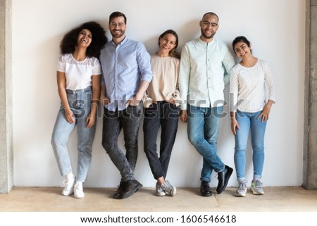 Portrait of happy millennial multiracial friends stand in row near white wall look at camera smiling, positive overjoyed diverse young people posing together for group picture, friendship concept