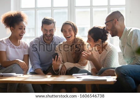 Happy excited international millennial friends sit at table laugh watching funny video on modern smartphone together, overjoyed multiethnic young people have fun smile at cellphone joke Royalty-Free Stock Photo #1606546738
