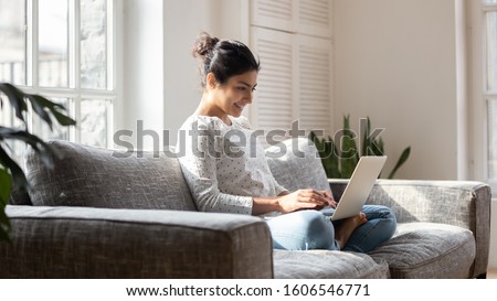 Smiling indian millennial girl sit relax on couch using modern laptop browsing unlimited wireless internet, happy young woman freelancer work on computer typing texting from home, technology concept Royalty-Free Stock Photo #1606546771