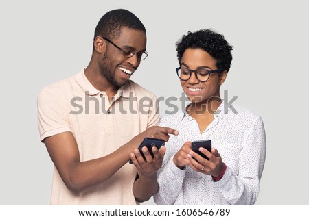 Attractive african ethnicity couple hold smart phones having fun in internet, guy help explain to girl new cool free online application, point finger show to photos, isolated on gray studio background