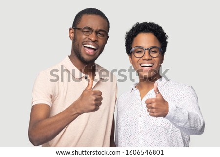 Overjoyed African couple in glasses showing thumbs up gesture looking at camera feels happy isolated on gray studio background, girl guy with wide white smile recommend services or products ad concept