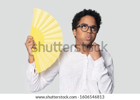 Mixed-race african girl rolled her eyes feels unwell too hot isolated on gray background holding yellow fan saves from unbearable heat fanning herself, lack of fresh air during hot weather concept Royalty-Free Stock Photo #1606546813