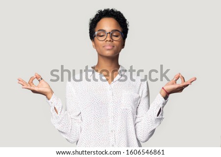 Pretty african woman wearing casual blouse and glasses keep calm feeling peaceful mood doing meditation pose isolated on grey background, no stress, anxiety relieve, self-control concept studio shot