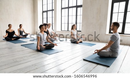 Instructor trainer teaching diverse young people at group lesson, doing Padmasana exercise, practicing yoga, sitting in Lotus pose on mats, working out, meditating in modern yoga studio Royalty-Free Stock Photo #1606546996