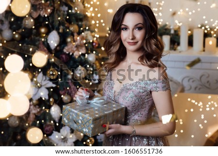 Happy new year. Woman with christmas box gift. Beauty portrait of young attractive girl wearing makeup in studio. Healthy hair style. Elegant lady in dress over christmas tree lights background