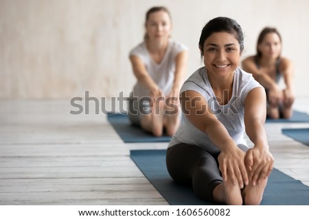 Smiling Indian woman practicing yoga at group lesson, doing Seated forward bend exercise, looking at camera, diverse people stretching in paschimottanasana pose on mats, portrait with copy space Royalty-Free Stock Photo #1606560082
