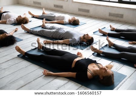Young sporty people with closed eyes meditating in Savasana pose on floor, practicing yoga at group lesson, doing Corpse exercise on mats, training, working out in modern yoga studio, center Royalty-Free Stock Photo #1606560157